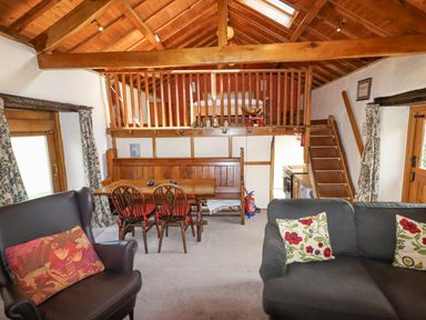 A living area with wooden dining table and chairs, a dark armchair, a sofa with floral cushions, wooden staircase and railing at The Shippon in Llechwedd The Conwy Valley