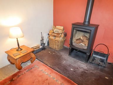 A wood burning stove with logs and a basket of firewood beside a wooden side table with a lamp at The Shippon in Llechwedd The Conwy Valley