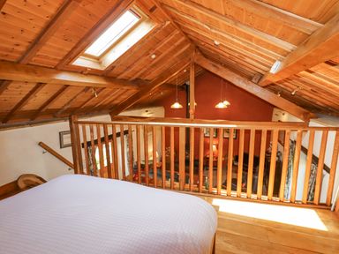 A loft bedroom with a wooden ceiling skylight and a wooden railing overlooking a living area with hanging lights at The Shippon in Llechwedd The Conwy Valley
