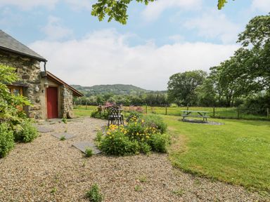 A stone building with a red door a gravel path flower beds metal chairs and a picnic table on grass with trees and hills in the background at The Shippon in Llechwedd The Conwy Valley