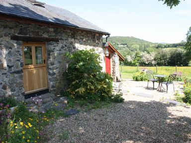 A stone cottage with a wooden door and a red door next to a garden with flowers and a metal table and chairs set outside at The Shippon in Llechwedd The Conwy Valley