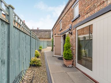 A narrow outdoor pathway with gravel and plants along a brick wall next to a green wooden fence at Upstairs Downstairs Cottage in Snainton near Scarborough
