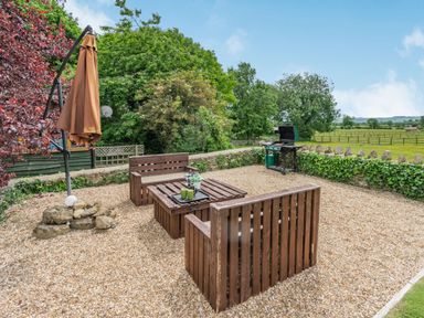 An outdoor patio area with wooden seating and table a closed umbrella and a barbecue grill surrounded by a stone wall and trees at Upstairs Downstairs Cottage in Snainton near Scarborough