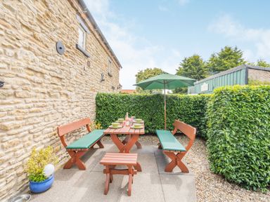 An outdoor patio area with wooden benches and table with green cushions and umbrella next to a stone wall and hedges at Upstairs Downstairs Cottage in Snainton near Scarborough