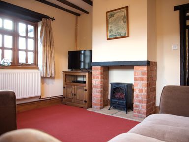 A living room with a fireplace a television on a wooden cabinet and a window with curtains at Yew Tree Cottage in Little Malvern near Malvern