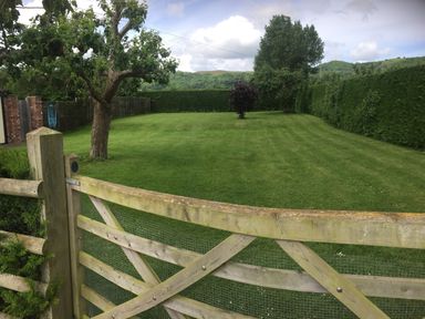 A garden with a lawn a wooden gate a tree and a hedge at Yew Tree Cottage in Little Malvern near Malvern