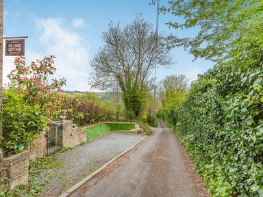 A narrow country lane with green hedges and a gravel driveway with a stone wall and gate at Hillside Cottage in Peasedown St John near Bath