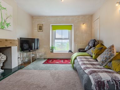 A living room with a sofa pillows a television on a glass stand and a window with a green blind at Hillside Cottage in Peasedown St John near Bath