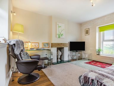 A sitting room with a leather armchair, glass table with pictures, fireplace, television, and window with green blind at Hillside Cottage in Peasedown St John near Bath
