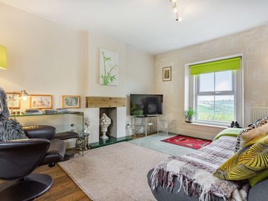 A living room with a sofa and pillows next to a window with green blind and a TV on a glass stand at Hillside Cottage in Peasedown St John near Bath