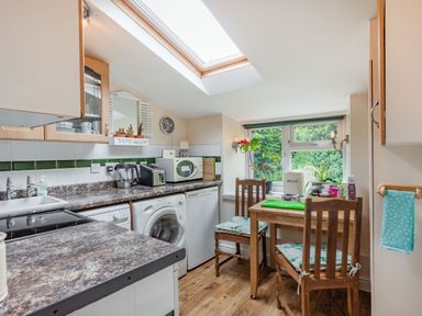 A kitchen with wooden chairs and table washing machine microwave and plants by the window at Hillside Cottage Peasedown St John near Bath