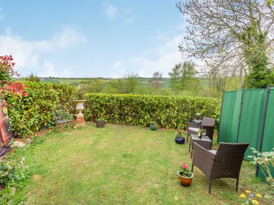 A garden with two wicker chairs and a small table with a bottle and glass surrounded by hedges at Hillside Cottage in Peasedown St John near Bath