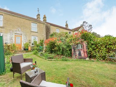 A garden with wicker chairs and a glass table in front of a stone house at Hillside Cottage in Peasedown St John near Bath