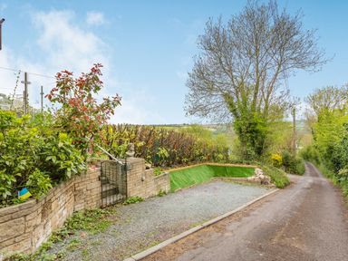 A narrow road next to a gravel parking area and greenery with a small gate and a stone bust at Hillside Cottage in Peasedown St John near Bath