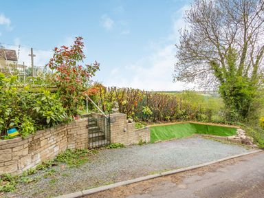 A gravel parking area with stone walls and a gate leading to a garden with plants and trees at Hillside Cottage in Peasedown St John near Bath