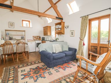 A room with wooden beams a blue sofa a rocking chair a patterned rug a dining table and a small kitchen at High Park Barn in Uffculme Blackdown Hills