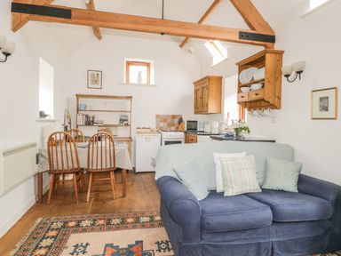 A living area with a blue sofa and pillows a dining table with wooden chairs and a kitchen with wooden cabinets and shelves at High Park Barn in Uffculme Blackdown Hills
