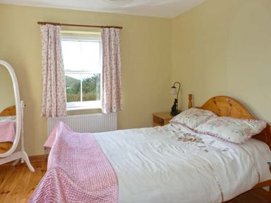 A bedroom with a wooden bed and bedside table a standing mirror and window with floral curtains at The Bay in Caherdaniel County Kerry