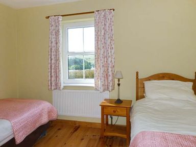 A bedroom with two single beds a wooden nightstand with a lamp and a window with floral curtains at The Bay in Caherdaniel County Kerry