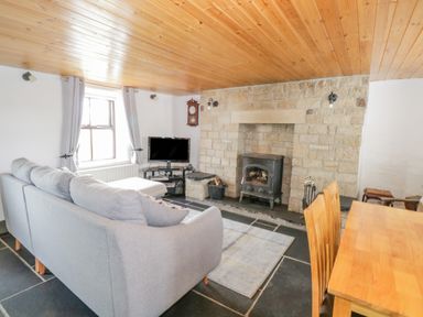 A living room with a gray sofa a wood stove a TV and a wooden dining table at Mary Kates Cottage in Narin County Donegal