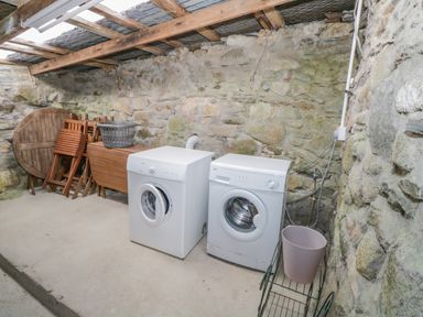 A laundry room with a washing machine and dryer against a stone wall with wooden furniture and a plastic basket at Mary Kates Cottage in Narin County Donegal