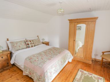 A bedroom with a bed floral quilt wooden wardrobe with mirror wooden nightstands lamps and wooden chair at Mary Kates Cottage in Narin County Donegal