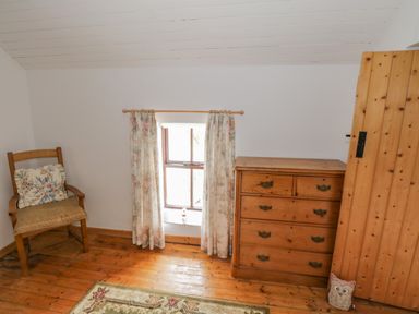 A small room with a wooden chair a floral cushion a window with floral curtains a wooden chest of drawers and a wooden door at Mary Kates Cottage in Narin County Donegal