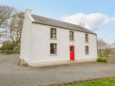 A white two-story house with a red door and four windows surrounded by trees at Mary Kate's Cottage in Narin County Donegal