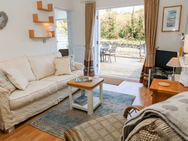 A living room with a cream sofa small coffee table television and open doors to a deck with outdoor chairs at Waterside Cottage in Little Petherick Creek near Padstow