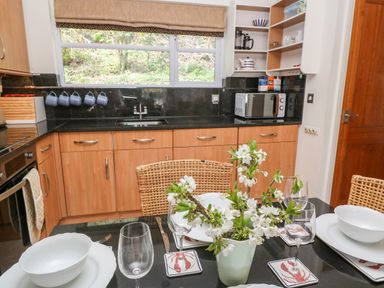 A kitchen with wooden cabinets a sink microwave and a dining table set with plates glasses and a flower vase at Waterside Cottage in Little Petherick Creek near Padstow