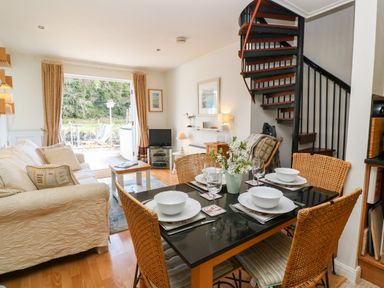A dining table set with bowls and wine glasses next to a living room with a sofa and spiral staircase at Waterside Cottage Little Petherick Creek near Padstow