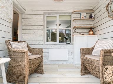 A sitting area with two wicker chairs and cushions in a room with whitewashed wooden walls and shelves at Waterside Cottage in Little Petherick Creek near Padstow