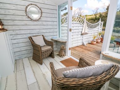 A sunroom with wicker chairs and striped cushions next to glass doors opening to a wooden deck at Waterside Cottage in Little Petherick Creek near Padstow