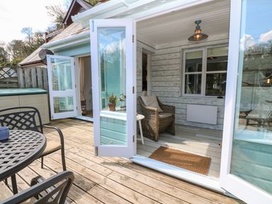 An outdoor wooden deck area with a round metal table and chairs next to a covered porch with a wicker armchair and open white framed glass doors at Waterside Cottage in Little Petherick Creek near Padstow