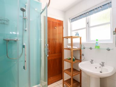 A bathroom with a glass shower enclosure a wooden door a wooden shelving unit and a white sink with a window above at Waterside Cottage in Little Petherick Creek near Padstow