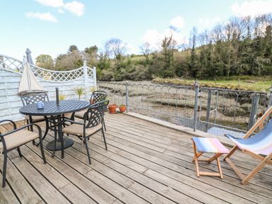 An outdoor wooden deck with a round metal table and four chairs near a railing by a creek at Waterside Cottage in Little Petherick Creek near Padstow