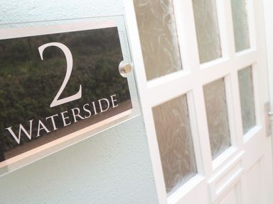 A black and silver house number sign with the number 2 and word waterside on a wall next to a white door with frosted glass panels at Waterside Cottage in Little Petherick Creek near Padstow