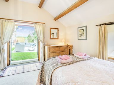 A bedroom with a bed, wooden dresser, lamp, framed pictures, curtains, and an open door leading to an outdoor seating area at Moresdale Bank Cottage Lambrigg near Sedbergh and Kendal