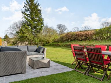 An outdoor patio with wicker sofa and table and wooden dining table with red cushioned chairs on grass at Moresdale Bank Cottage Lambrigg near Sedbergh and Kendal