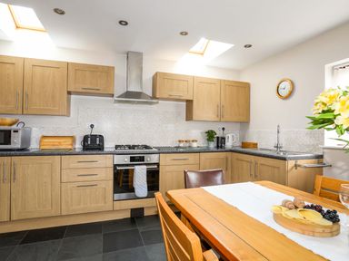 A kitchen with wooden cabinets, a black countertop, a gas stove, microwave, toaster, kettle, a dining table with chairs and food at Penrallt Cottage in Y Felinheli near Caernarfon