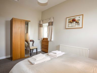 A bedroom with a bed and folded towels on it a wooden wardrobe with a mirror a chair a chest of drawers and a framed flower picture on the wall at Penrallt Cottage in Y Felinheli near Caernarfon