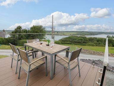 An outdoor deck with a table and four chairs overlooking a river and green landscape at Penrallt Cottage in Y Felinheli near Caernarfon