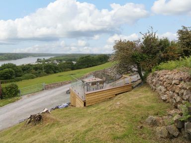 An outdoor wooden deck with a glass railing and table with chairs overlooking a rural countryside and river at Penrallt Cottage in Y Felinheli near Caernarfon