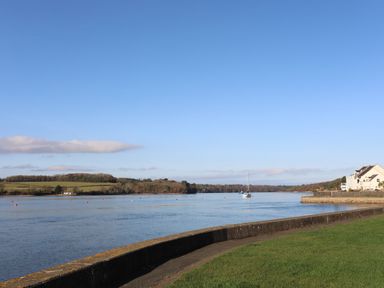 A river with a sailboat and houses on the shore near a curved concrete wall and grass at Penrallt Cottage in Y Felinheli near Caernarfon