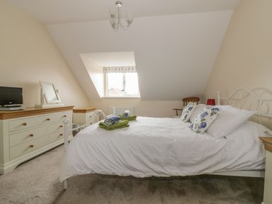 A bedroom with a bed with white bedding and floral pillows a dresser with a mirror and television and a window at Sandlings in Hollesley near Woodbridge