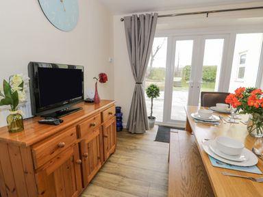 A dining room with a wooden sideboard holding a television and flowers and a dining table set with dishes and flowers at Gwelfor in Morfa Bychan