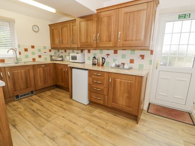 A kitchen with wooden cabinets a small fridge microwave kettle toaster and a white door with a fire exit sign at Gwelfor Morfa Bychan