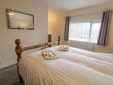 A bedroom with a wooden bed frame white bedding folded towels on the bed a window with vertical blinds and curtains a mirror on the wall at Gwelfor in Morfa Bychan