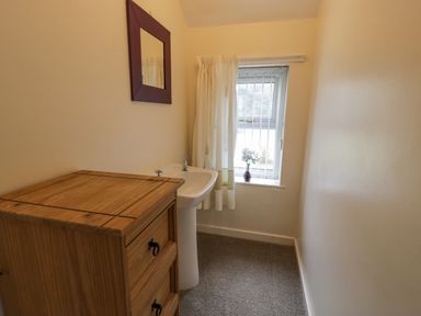 A small room with a wooden chest of drawers a white pedestal sink a window with vertical blinds and a vase of flowers and a wall mirror at Gwelfor in Morfa Bychan