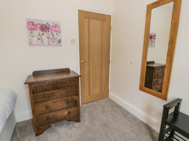 A wooden chest of drawers under a flower painting next to a wooden door with a mirror and a small black chair in a carpeted room at Gwelfor in Morfa Bychan